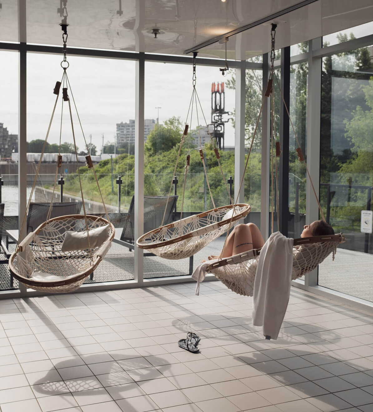 Espace détente intérieur avec fauteuils suspendus en rotin donnant sur le fleuve, une personne relaxe dans un hamac-balançoire. / Indoor relaxation area with hanging rattan chairs overlooking the river, one person resting in a hammock swing.