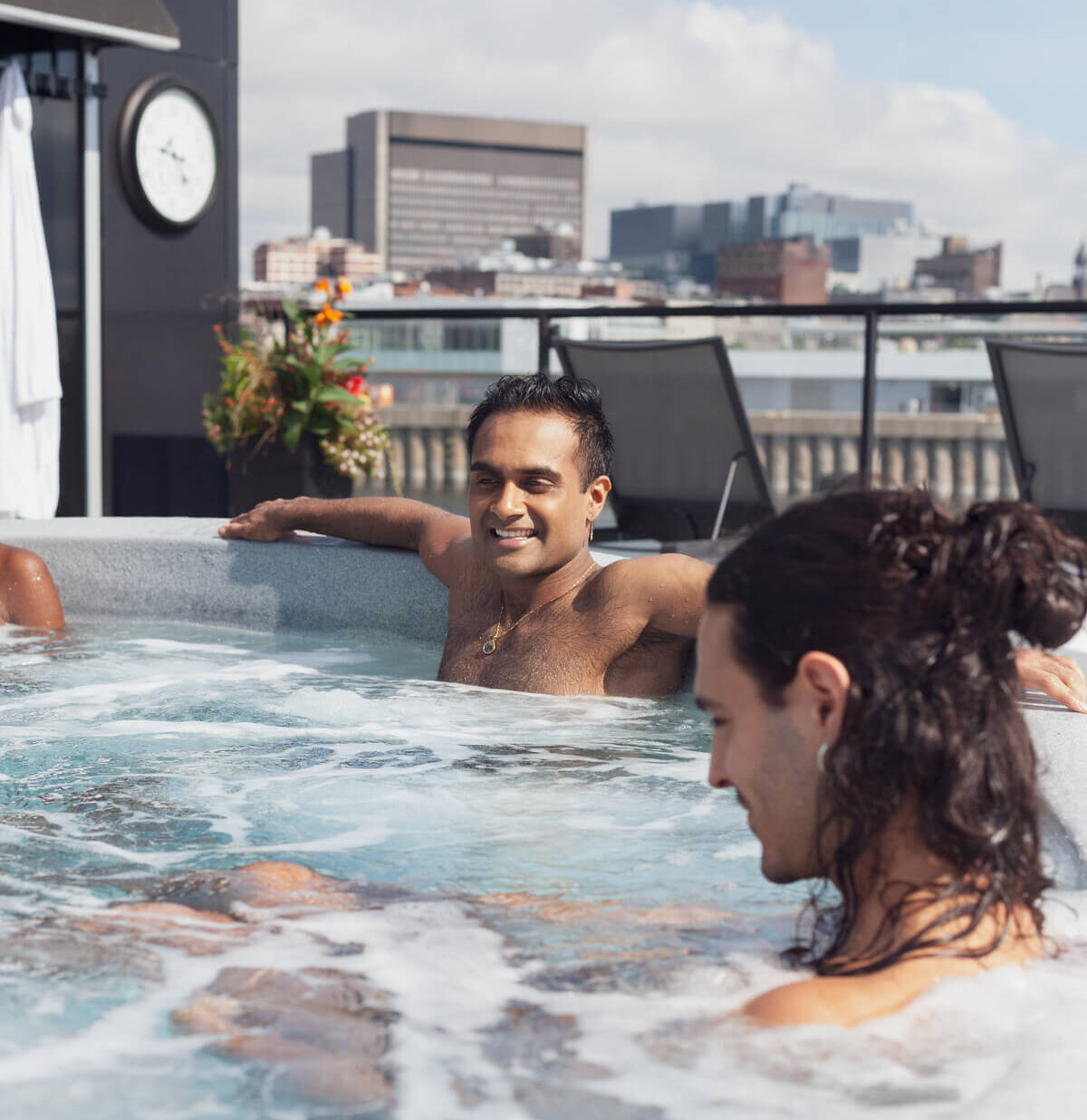 Hommes détendus dans un jacuzzi extérieur, profitant de la vue sur la ville de Montréal. / Men relaxing in an outdoor hot tub, enjoying the view of Montreal city.
